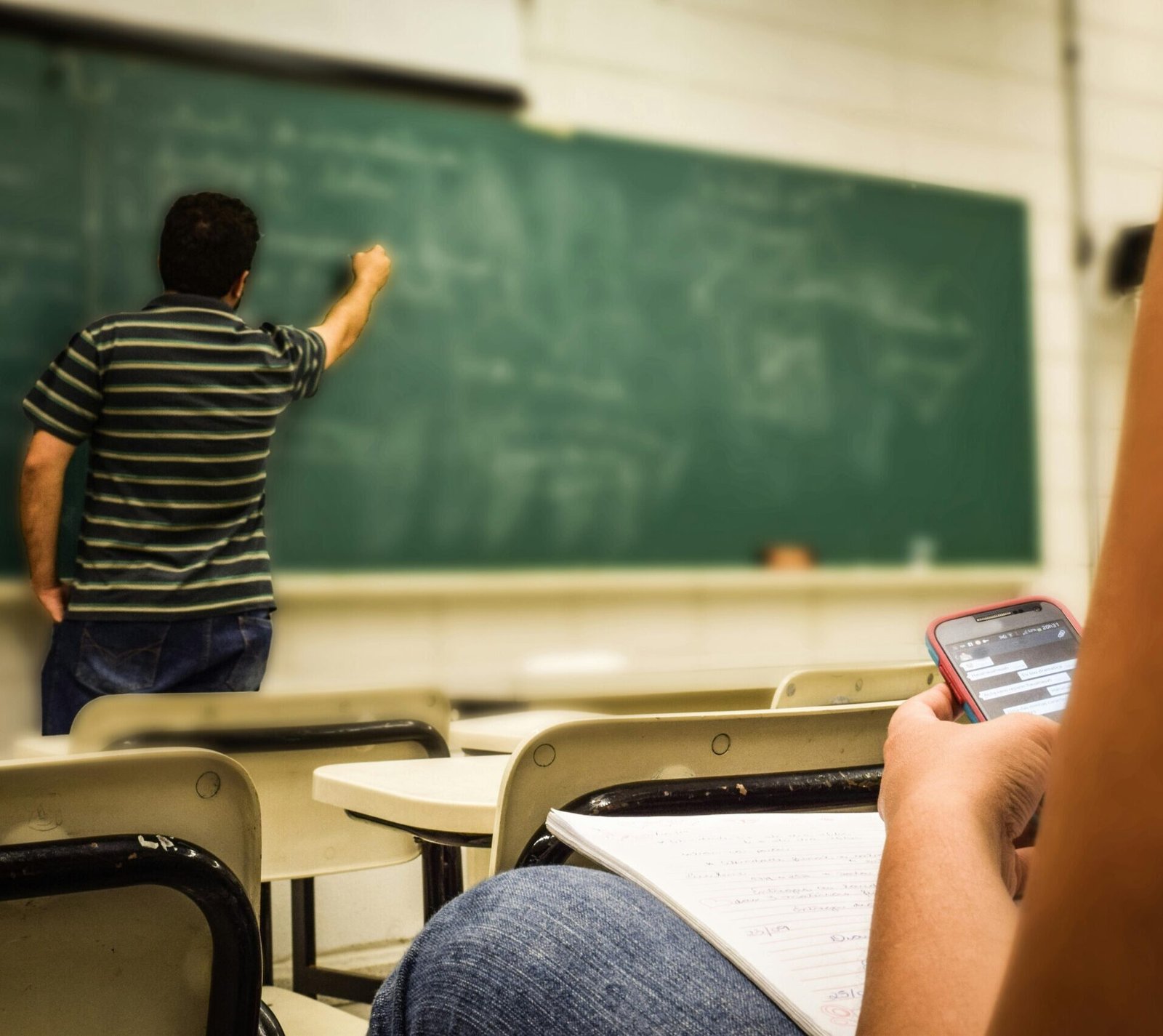 Student texting in a classroom while teacher is writing on the blackboard.